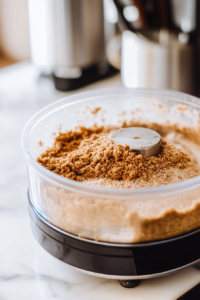A close-up shot of a glass food processor on a white marble cooktop, processing graham crackers into fine crumbs. A mixture of brown sugar is added, and the machine is whirring, creating a light dusting of crumbs.