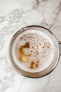 A saucepan on the stovetop with melted butter, brown sugar, and heavy cream bubbling together to create salted butterscotch sauce. A wooden spoon rests on the side, and a jar is ready for storage