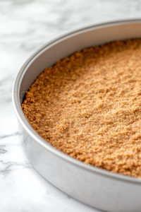 A 9-inch springform pan with a pressed crust made from crushed graham crackers and butter, ready to be baked. The pan sits on a parchment-lined baking sheet.