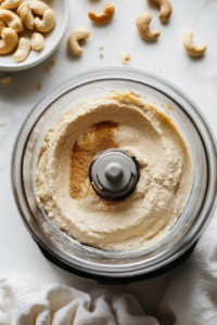 Action shot of cashews, tofu, nutritional yeast, lemon juice, and spices being blended together in a food processor. The creamy ricotta mixture is smooth, with a few cashews on the countertop for context.