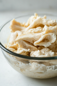 Side view of oyster mushrooms soaking in a vegan buttermilk mixture inside a glass bowl. The mushrooms are fully coated, and a colorful kitchen towel is draped beside the bowl on the counter.