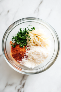 A shallow bowl on a white marble cooktop with grated parmesan cheese, potato starch, paprika, and dried parsley being whisked together with a fork until well combined