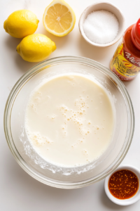 Close-up of a large bowl with almond milk and lemon juice being whisked into a vegan buttermilk mixture. The mixture looks slightly curdled, with a small bowl of coconut sugar and a bottle of hot sauce nearby on the counter.