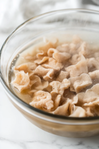 Close-up of soy curls soaking in hot vegan broth in a glass bowl. Steam rises gently from the mixture, and a spoon rests beside the bowl, creating a warm and cozy kitchen atmosphere.