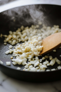 Close-up shot of a black skillet on the white marble cooktop, sizzling olive oil with chopped onion and crushed garlic. The onion is softening, and steam rises from the pan. A wooden spoon rests beside, ready for stirring.