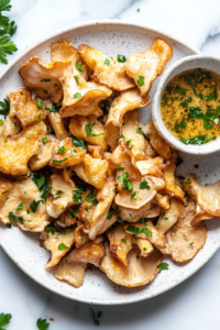 A serving plate of golden-fried oyster mushrooms arranged neatly, garnished with fresh parsley. A small bowl of dipping sauce sits beside the plate on a rustic table setting with a napkin, fork, and a glass of lemonade.