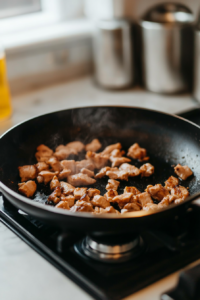 Close-up shot of chopped chicken in a sizzling black non-stick skillet on the white marble cooktop. The chicken is mostly cooked and turning golden brown.