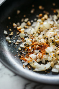 Close-up shot of a black skillet on the white marble cooktop, with ground ginger, coriander, smoked paprika, black pepper, salt, and maple syrup being stirred into sautéed onions and garlic. The spices release a fragrant cloud as they toast.