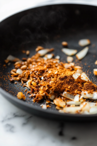 Close-up shot of a black skillet on the white marble cooktop, with curry powder, cumin, and cinnamon being stirred into the sautéed onions and garlic. The spices are toasting and releasing a fragrant aroma.