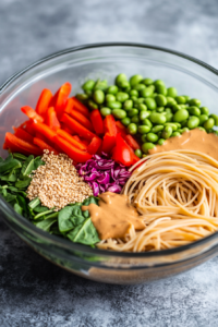 A large serving bowl with cooked whole wheat spaghetti, shredded cabbage, red bell pepper slices, spinach, edamame, and chopped cilantro, with the peanut sauce being poured over the top.