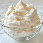 Close-up shot of the fluffy and smooth whipped aquafaba in the glass bowl, garnished with a dollop on a spoon beside it. The bowl sits on the white marble cooktop, with light reflecting off the creamy texture