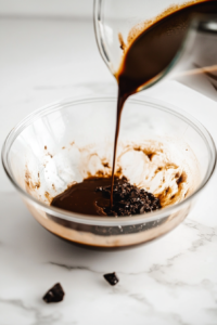 The same glass mixing bowl on a white marble cooktop, now with melted coconut oil, plant-based milk, and vanilla extract being poured in. The batter starts to come together, looking rich and chocolatey.