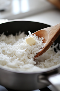 Shredded coconut, butter, and vanilla are stirred into the thickened coconut filling in a saucepan on the white marble cooktop. The mixture is rich and creamy.