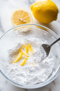 Close-up shot of the glass mixing bowl on the white marble cooktop, now with powdered sugar, lemon juice, lemon zest, and vanilla added to the whipped cream cheese. A spatula scrapes the sides as everything mixes to a silky consistency. --ar 4:6