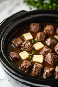 A close-up of a black slow cooker on a white marble cooktop, as rich beef broth is poured over the seasoned steak bites. Small pats of butter are placed on top, beginning to melt and infuse the meat with extra flavor.