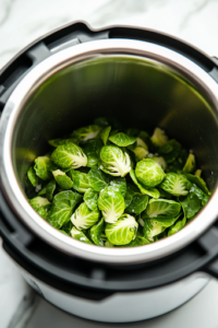 A close-up shot of fresh Brussels sprouts being poured into the Instant Pot on a white marble cooktop. The vibrant green sprouts are coated with garlic, salt, and cracked pepper, glistening as they absorb the flavorful broth.