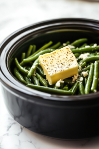 A close-up shot of a stick of butter cut into chunks, evenly placed on top of the green beans and seasonings inside the black slow cooker on the white marble cooktop. The butter will melt slowly, infusing the dish with a creamy, rich taste.