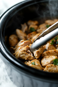 A close-up shot of the black crockpot on the white marble cooktop, with reduced sodium chicken broth being poured over the ingredients. The liquid blends with the soups and seasonings, creating a creamy and aromatic base for the slow-cooked chicken.