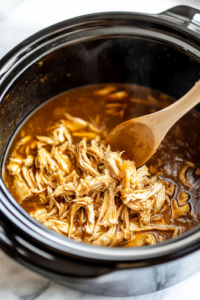 A close-up shot of a black slow cooker on the white marble cooktop, as the freshly shredded chicken is added back into the rich, golden broth. A wooden spoon stirs gently, ensuring the chicken is evenly distributed throughout the soup.