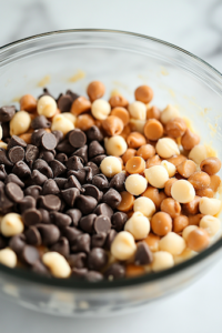 A close-up shot of chocolate chips and small caramel candy pieces being folded into the cookie dough in the glass mixing bowl on the white marble cooktop. The dough looks rich and textured, with generous chunks of mix-ins spread throughout.