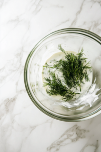 A close-up shot of a small glass mixing bowl on the white marble cooktop, with sour cream (or Greek yogurt), mayonnaise, freshly chopped dill, white vinegar, and granulated sugar being added, preparing the base for a creamy and flavorful dressing.