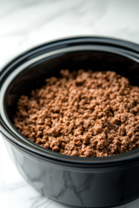 A close-up shot of a black slow cooker on a white marble cooktop, now filled with freshly browned ground beef. The beef is evenly crumbled and spread across the bottom of the slow cooker, ready to absorb the rich flavors of the upcoming ingredients.