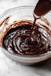 A close-up shot of boiling water (or hot coffee) being poured into the chocolate cake batter in the glass mixing bowl on the white marble cooktop. A spatula stirs the mixture, making it smooth and glossy as the liquid enhances the chocolate flavor.
