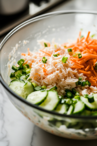 A close-up shot of a large glass mixing bowl on the white marble cooktop, with shredded imitation crab, julienned cucumber, and carrot being combined with the freshly prepared creamy dressing, ready to be mixed into a flavorful kani salad.