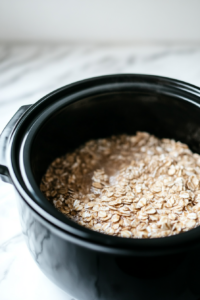 A black slow cooker on the white marble cooktop, with steel-cut oats and water being poured in. The ingredients mix together, ready to cook overnight.