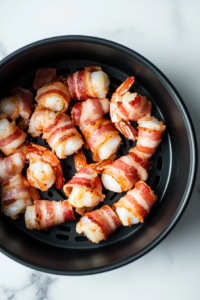 Close-up shot of the air fryer basket on the white marble cooktop, neatly arranged with bacon-wrapped shrimp placed seam-side down. The shrimp glisten slightly, ready for cooking.