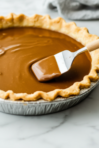 Close-up shot of the smooth chocolate filling being poured into the golden-baked pie crust on the white marble cooktop. A spatula spreads the mixture evenly before placing it into the refrigerator to chill.