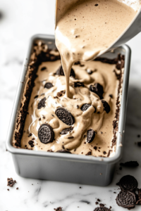 Close-up shot of the cheesecake mixture being poured over the Oreo crust in the loaf pan. The cheesecake is ready to be baked, sitting on the marble countertop