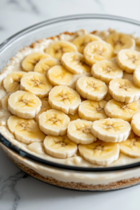 A layer of sliced bananas is evenly placed over the crust. The pudding-coconut mixture is spread on top, creating a smooth finish