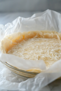 A glass pie plate is about to be inverted onto the dough, sitting on the white marble cooktop. The parchment paper is being peeled off, and the edges of the dough are being trimmed to fit the plate.