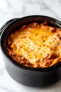A close-up shot of the black slow cooker on the white marble cooktop, fully assembled with alternating layers of meat sauce, noodles, and cheese. The top layer is covered with golden mozzarella, and the lid is on, allowing the lasagna to slow-cook to perfection.