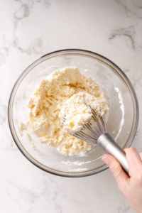 Close-up shot of softened cream cheese being whipped in a mixing bowl with an electric hand mixer until smooth and creamy