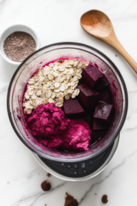 A high-speed blender filled with cooked beets, rolled oats, almond milk, cacao powder, vanilla extract, flaxseed, salt, baking powder, maple syrup, and beet juice, ready to be blended into a smooth batter.