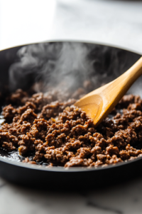 A black skillet on a white marble cooktop with lean ground beef sizzling and browning evenly. A wooden spatula is seen stirring the meat, breaking it into crumbles as steam rises, indicating the cooking process.