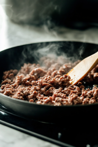 A close-up shot of a black cast-iron skillet on a white marble cooktop, where ground beef is sizzling and browning over medium heat. A wooden spatula stirs the crumbled beef, with small wisps of steam rising. The texture gradually changes from raw pink to a rich golden brown as excess grease pools in the pan, creating a savory aroma.