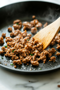 A close-up shot of a black skillet on a white marble cooktop, where ground sweet Italian sausage is sizzling to a perfect golden brown. A wooden spatula stirs the meat, releasing savory aromas, while small, crispy bits form on the pan, adding depth of flavor.