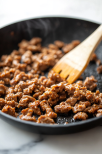 A black skillet on a white marble cooktop, with crumbled maple-flavored sausage sizzling and browning. A wooden spatula stirs the sausage as it cooks