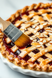 A pastry brush applies a light coat of dairy-free cream (with a touch of turmeric) over the lattice crust. Slivered almonds are being generously sprinkled on top.