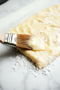 A pastry brush spreading melted butter over the crescent roll dough, with sugar being sprinkled evenly on top.