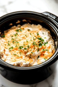A black crockpot on a white marble cooktop, filled with drained chunk chicken breast, softened cream cheese, shredded Colby Jack cheese, Buffalo sauce, and ranch dressing. The ingredients are placed together before mixing, ready to transform into a creamy, flavorful dip.