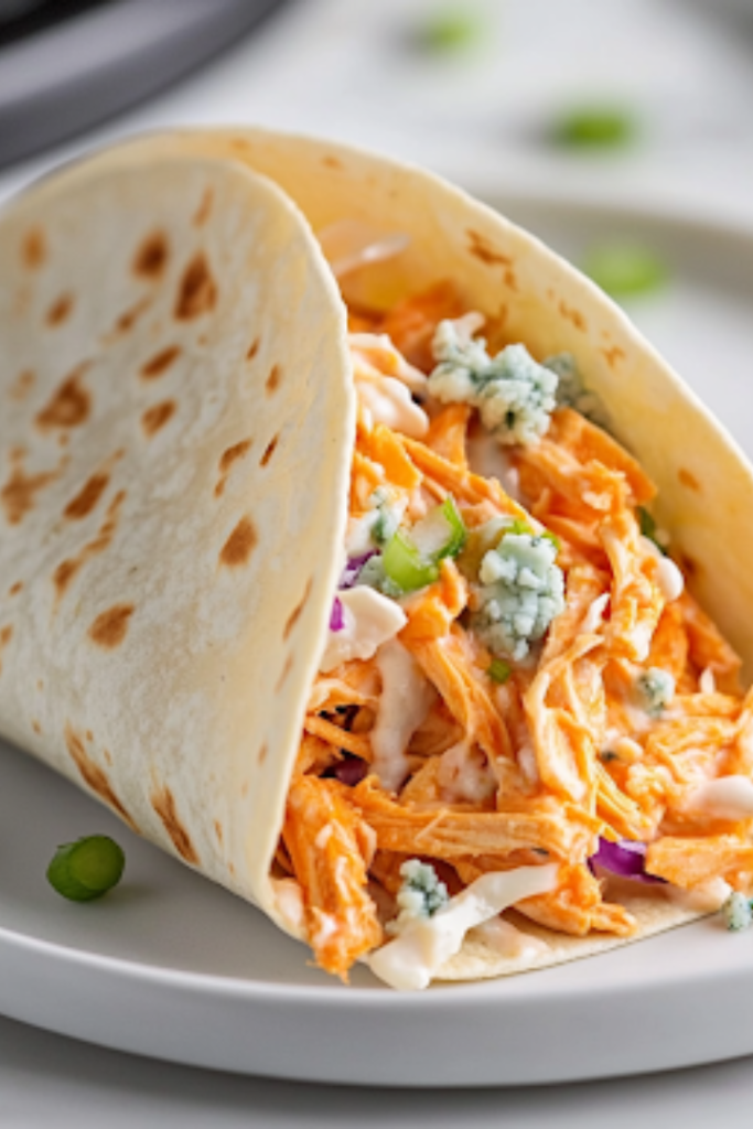 A freshly assembled Buffalo chicken taco sits on a white plate over the white marble cooktop. The warm tortilla is filled with spicy Buffalo chicken, creamy coleslaw, crumbled blue cheese, and sliced green onions. A small bowl of ranch dressing is placed beside it for dipping, completing the delicious dish.