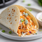 A freshly assembled Buffalo chicken taco sits on a white plate over the white marble cooktop. The warm tortilla is filled with spicy Buffalo chicken, creamy coleslaw, crumbled blue cheese, and sliced green onions. A small bowl of ranch dressing is placed beside it for dipping, completing the delicious dish.