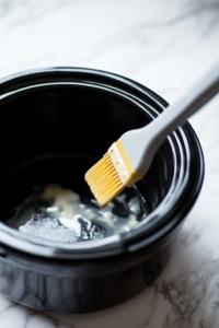 A close-up shot of a black slow cooker on the white marble cooktop, with a silicone brush spreading melted butter evenly on the interior surface. The slow cooker is being coated to prevent sticking and enhance flavor.