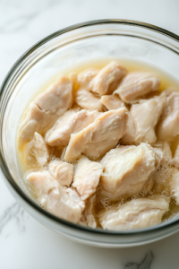 A glass bowl on a white marble cooktop, filled with fresh chicken pieces fully submerged in creamy buttermilk. Small bubbles form on the surface, indicating the marination process.