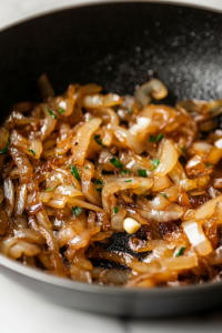A close-up shot of a black cast-iron skillet on a rustic wooden countertop, filled with thinly sliced yellow onions slowly caramelizing in melted butter. The onions glisten as they turn a deep golden brown, while a wooden spoon stirs them gently. Soft wisps of steam rise from the pan, enhancing the warm, savory aroma.