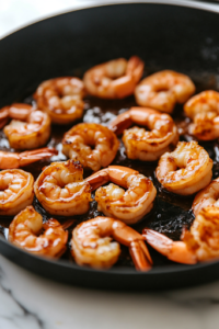 A close-up shot of the shrimp caramelizing in the black skillet over a white marble cooktop. The edges turn golden brown, and the sauce thickens into a glossy glaze, coating the shrimp evenly for a rich, flavorful finish.
